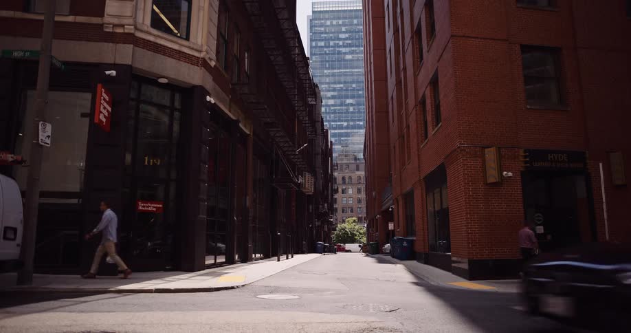 Pedestrians and a slow-moving car cross a sunny intersection in a city alley framed by classic brick buildings and a modern skyscraper