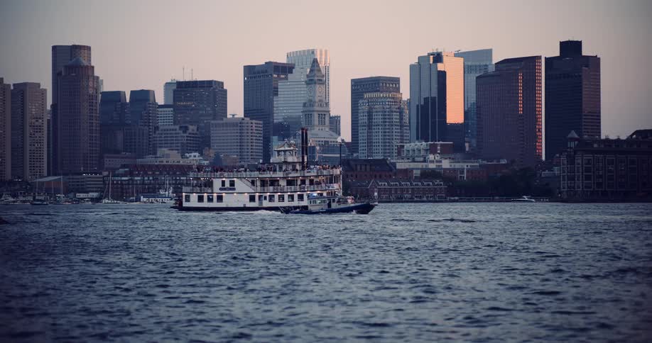A majestic riverboat tour cruises across Boston Harbor with the iconic city skyline reflecting the warm light of sunset in the background