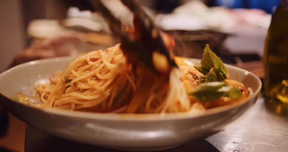 A delicious, steaming bowl of spaghetti with tomato sauce and basil is carefully plated with tongs in slow motion