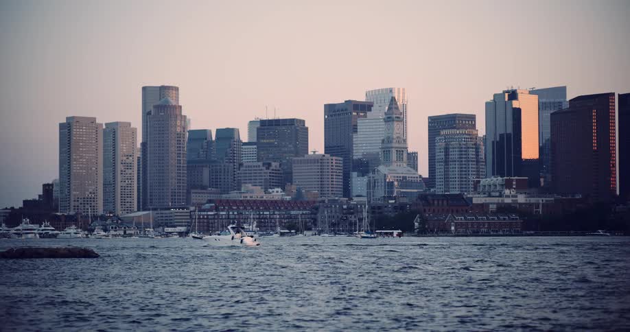 A motorboat sails through the Boston harbor at dusk with the city's iconic skyline in the background