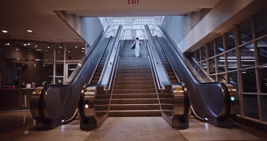 A woman in a white dress walks up a stationary escalator toward a brightly lit exit