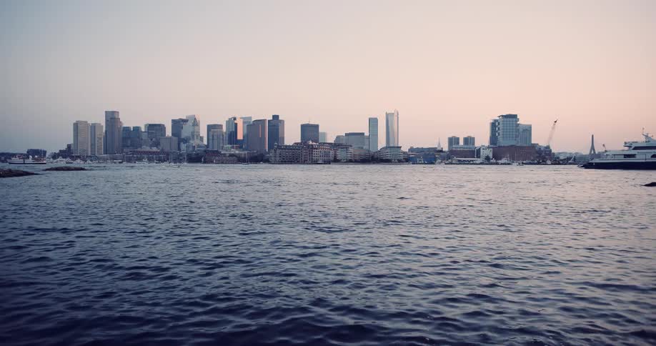 A large ferry sits docked in the Boston harbor with the city skyline in the background during a peaceful sunset