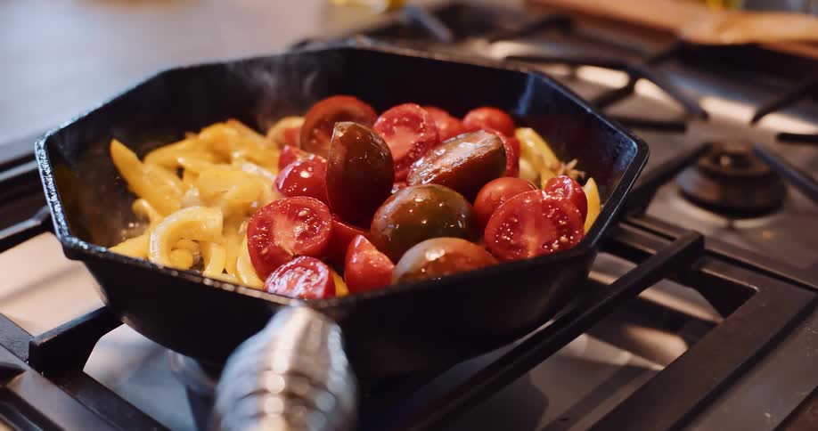A chef sautés yellow peppers and garlic before adding fresh heirloom tomatoes to a cast-iron skillet on a gas stove in slow motion