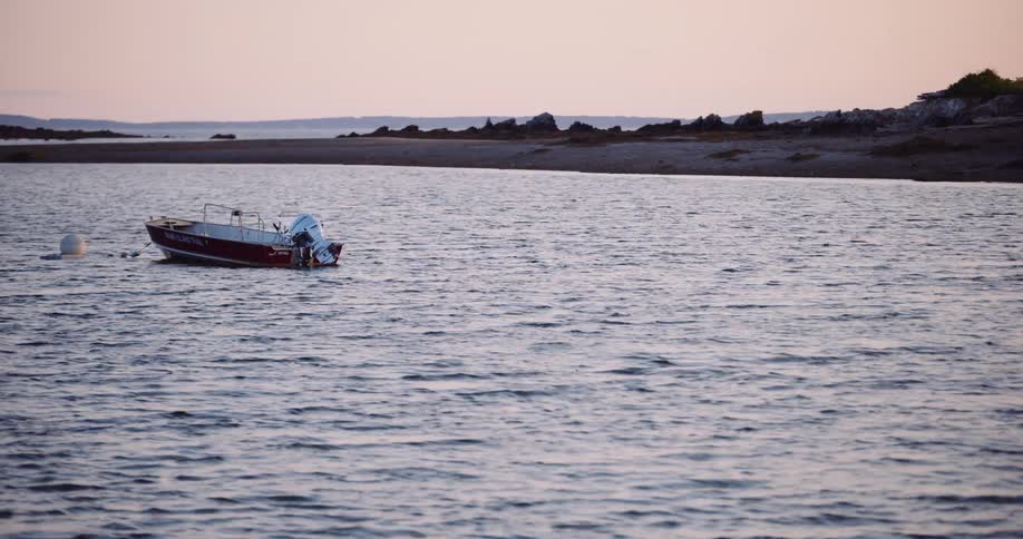 A single motorboat gently bobs in a quiet cove as the sun sets over a rocky shoreline