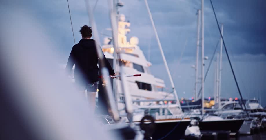 A man in a black hoodie stands on the deck of a boat in a marina at dusk, gazing at the illuminated city skyline