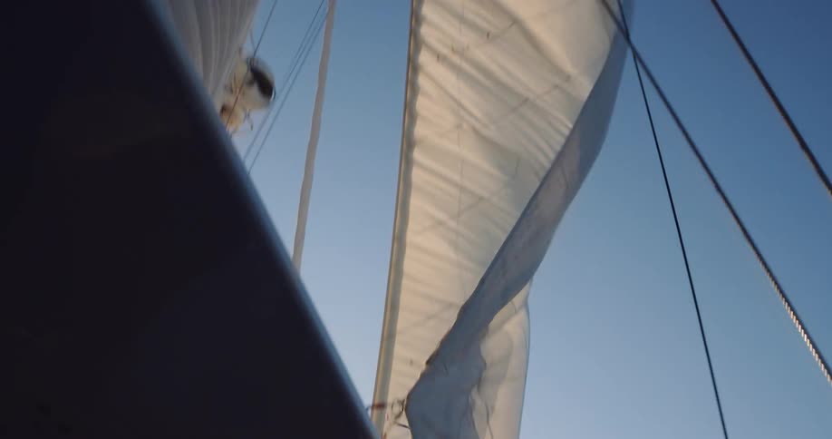 Looking up from the deck of a sailboat, the white sails gracefully billow in the wind against a brilliant blue sky