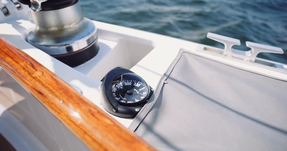 A close-up of a sailboat compass next to a winch, rocking back and forth while sailing on the water as shadows pass overhead