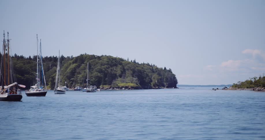 A slow pan reveals a tranquil ocean bay with sailboats anchored near a lush, tree-covered island on a beautiful sunny day
