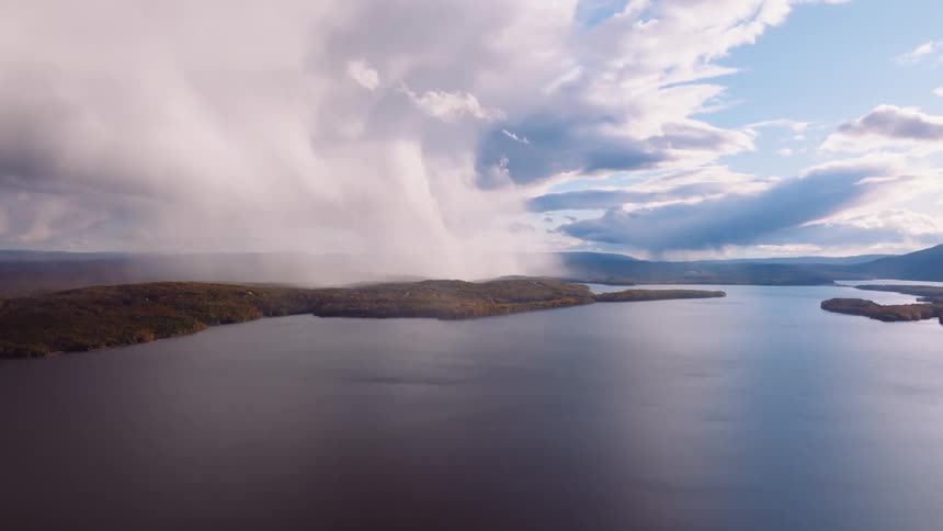 A stunning aerial view captures a dramatic, isolated rainstorm pouring from a low cloud onto a forested peninsula in a vast, calm lake during autumn