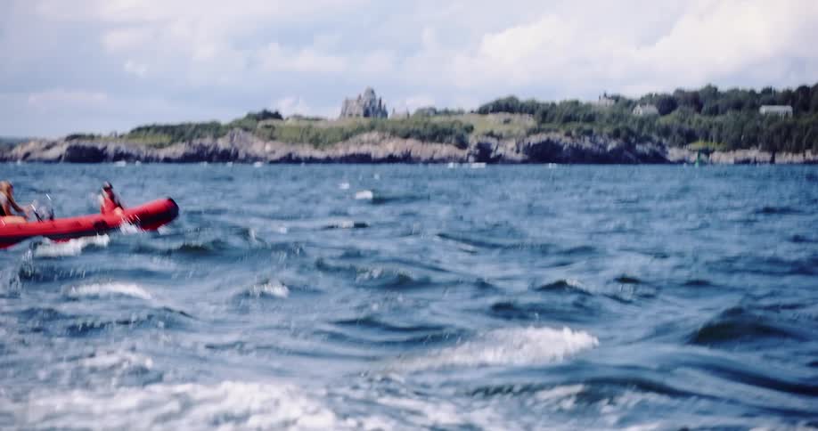 Two people navigate a red inflatable boat across choppy blue water with a majestic coastal mansion on the rocky shore in the distance