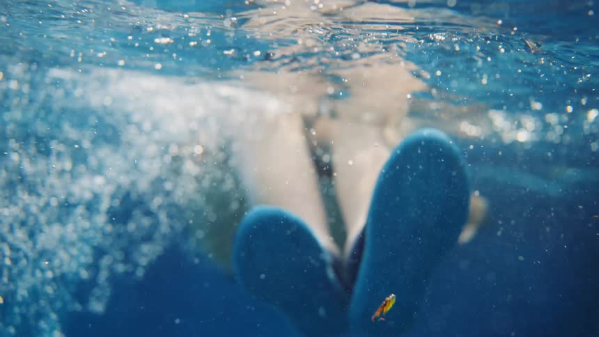 An underwater slow-motion view shows a pregnant woman in a bikini and water shoes floating peacefully on her back in a swimming pool