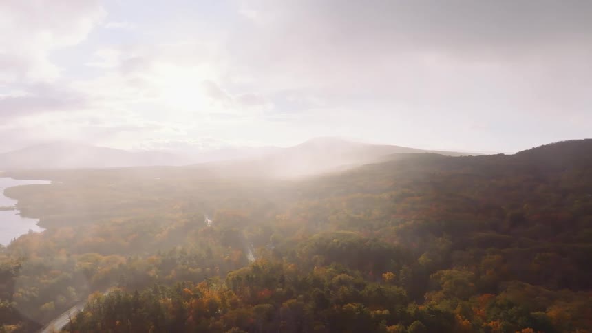 An ethereal aerial view captures the morning mist and sun rays illuminating a vast autumn forest landscape with mountains and a lake