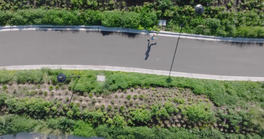 Aerial top-down view of a woman jogging along a winding paved path surrounded by lush green landscaping on a sunny day