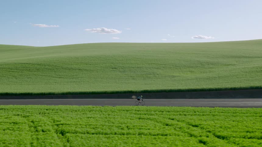 Parallel traverse alongside a man riding a bike on a paved road through picturesque rolling green hills