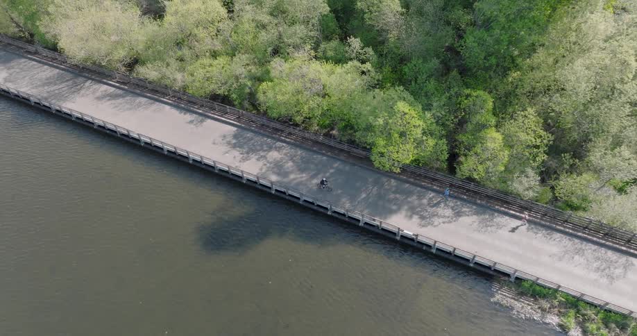 An aerial top-down view of a person cycling along a scenic path that runs between a tranquil river and a dense green forest