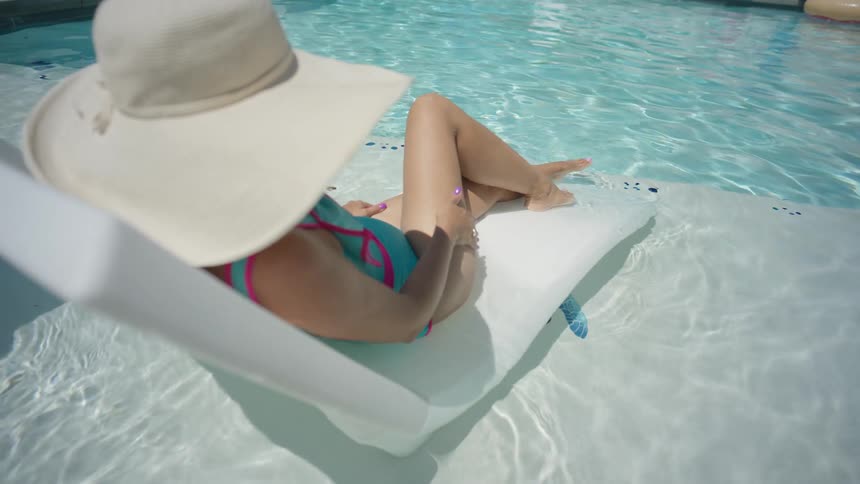 A woman in a big white hat relaxes on a lounge chair in the shallow end of a sparkling swimming pool on a sunny day