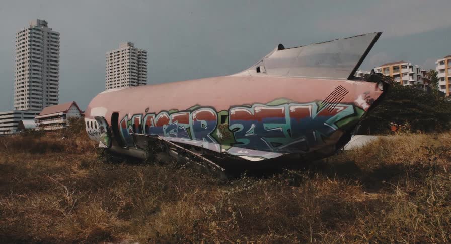 A wrecked airplane fuselage covered in graffiti sits abandoned in an overgrown urban field with city buildings in the background