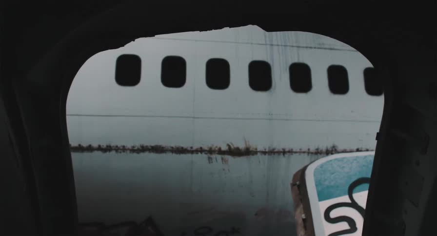 A view from inside a derelict airplane looks out through a broken, silhouetted window at another abandoned aircraft covered in rust and graffiti