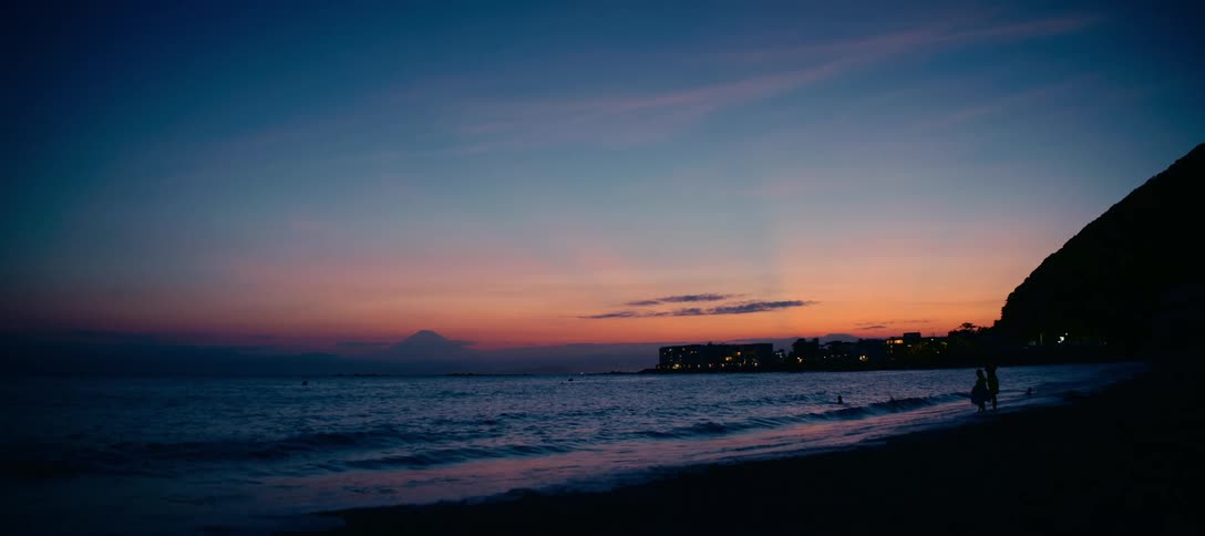 A serene view of a Japanese beach at dusk, with gentle waves lapping the shore and the silhouette of Mount Fuji against a colorful sunset sky