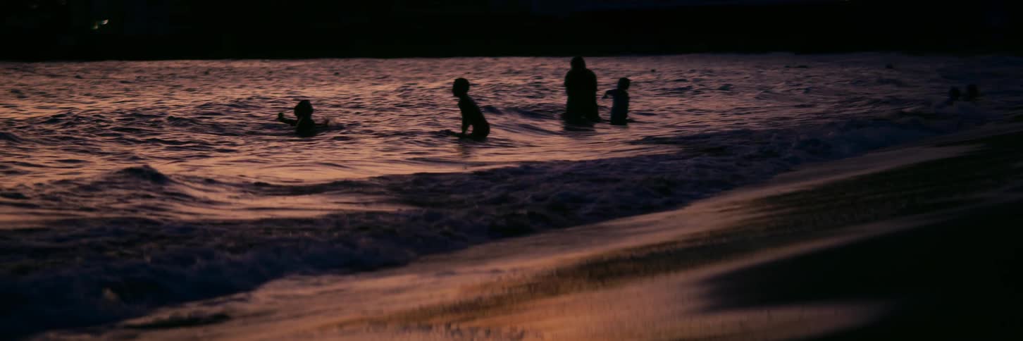 Silhouettes of children playing in the gentle ocean waves during a warm sunset in slow motion