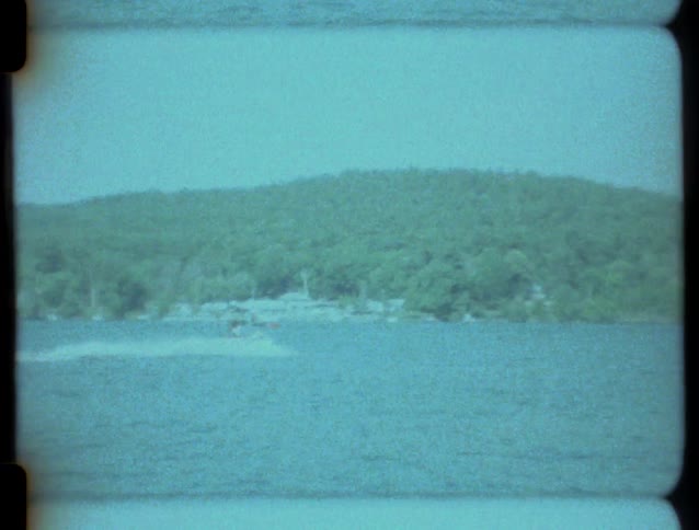 A grainy home movie captures a speedboat racing across a large lake with a tree-covered hill in the background