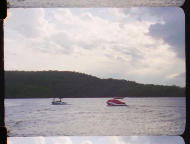 A vintage film shot captures two motorboats cruising across a lake on a cloudy summer day