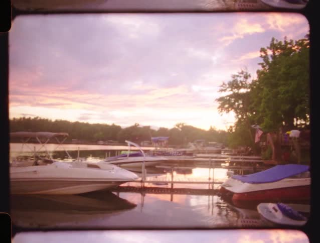 A vintage film pan of a serene lake dock with boats, revealing a beautiful sunset over the water