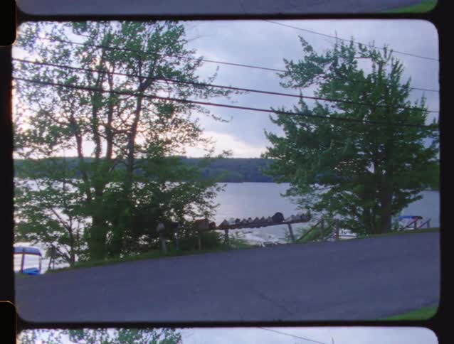 A car drives on a quiet lakeside road as the camera pans to boats on the water, captured on vintage 16mm film