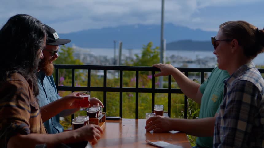 A group of friends enjoys a craft beer tasting, laughing and toasting on a scenic outdoor patio with a marina and mountains in the background