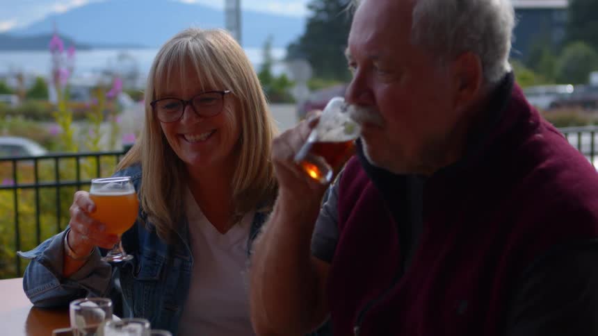 A happy senior couple laughs and enjoys a craft beer tasting together on a scenic outdoor patio overlooking the water