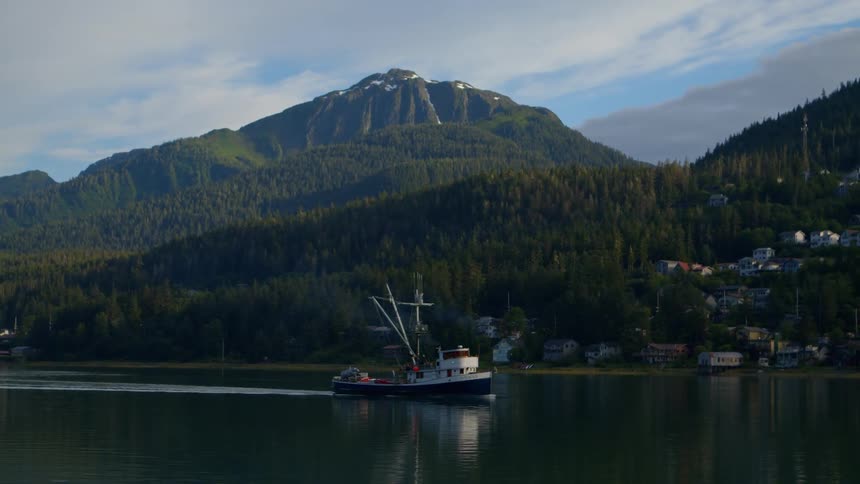 A fishing boat sails through a tranquil bay with a picturesque mountain and a forested village in the background