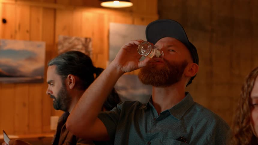 A smiling man with a red beard and backwards cap enjoys a drink from a small taster glass at a rustic bar