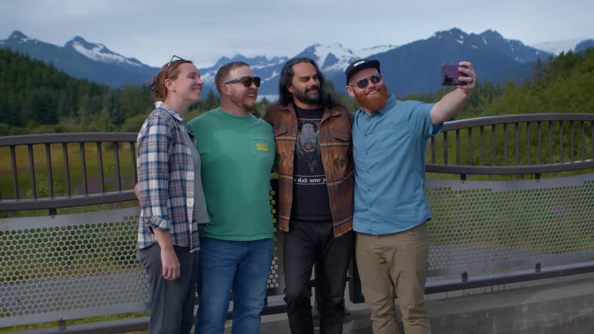 A group of happy friends take a selfie together on a bridge with a beautiful snow-capped mountain range in the background