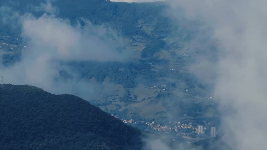 A slow zoom-out revealing a town nestled in a lush mountain valley through a blanket of moving clouds