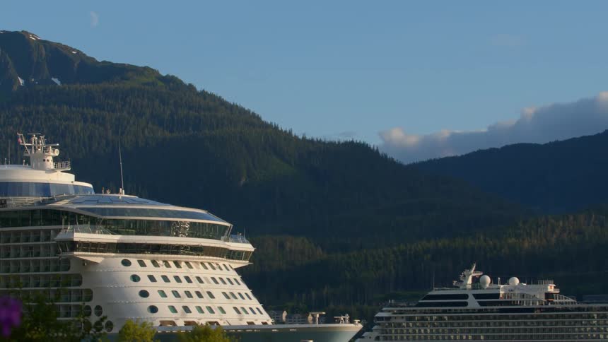 A large cruise ship is docked in a scenic harbor with a backdrop of lush, forested mountains and a crescent moon in the blue sky