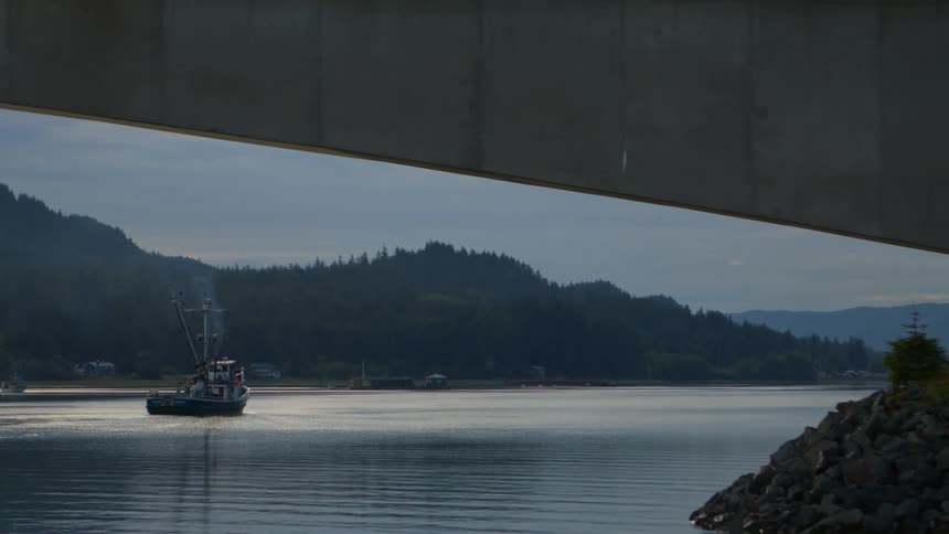 A fishing boat motors peacefully on calm water, framed by the underside of a bridge, with tree-covered hills in the background