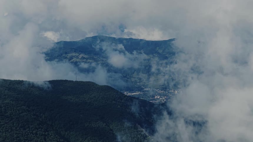 Thick clouds slowly drift over lush green mountains, revealing a small city hidden in the valley below