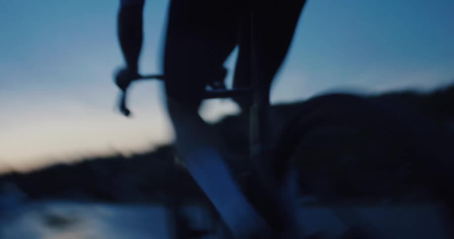 A blurry, artistic low-angle shot follows a cyclist's legs as they pedal hard on a road at dusk