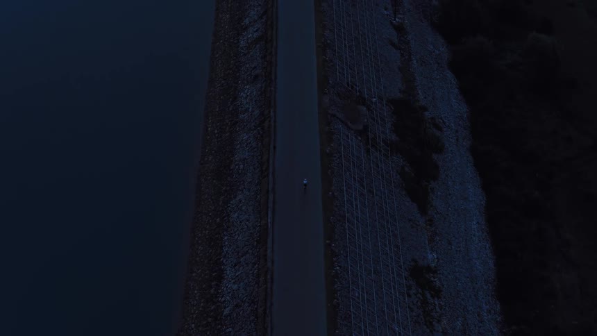 A dramatic top-down aerial shot follows a lone cyclist riding along a dark, empty road on top of a dam at night