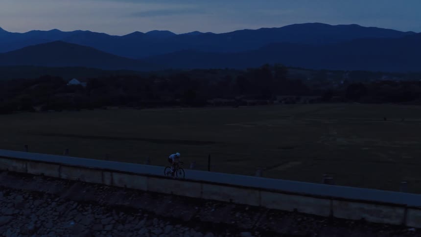 A cyclist rides on a dam road next to a tranquil lake at dusk, with mountains in the background