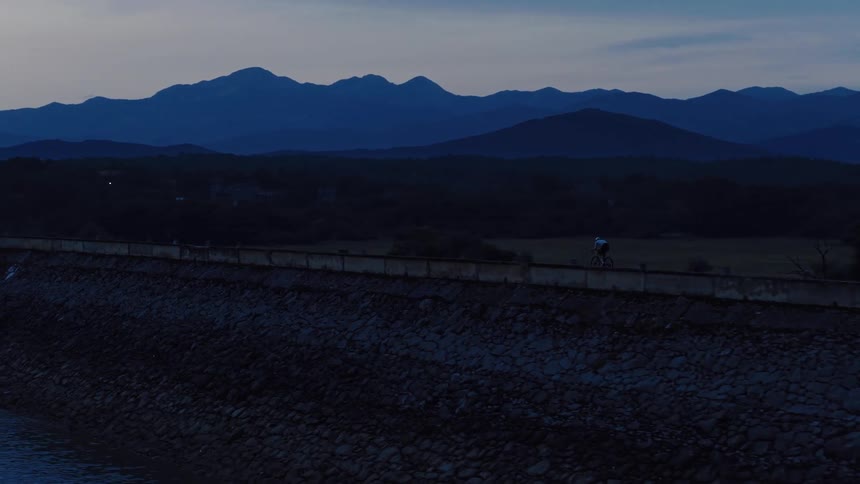 A lone cyclist rides across a remote dam road at twilight with mountains silhouetted in the background