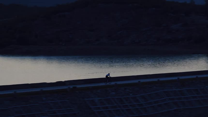 A solitary cyclist rides along a dark, moody lakeside road during the blue hour