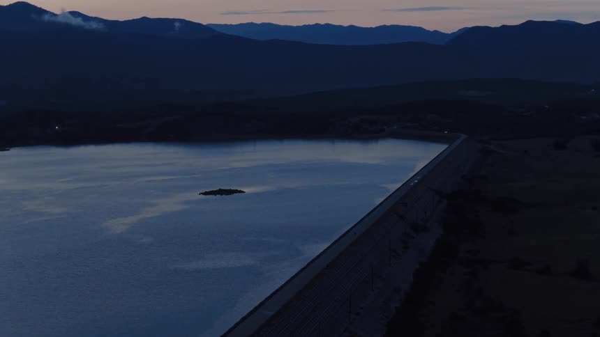 A serene aerial view shows a large reservoir and dam at twilight with silhouetted mountains in the background