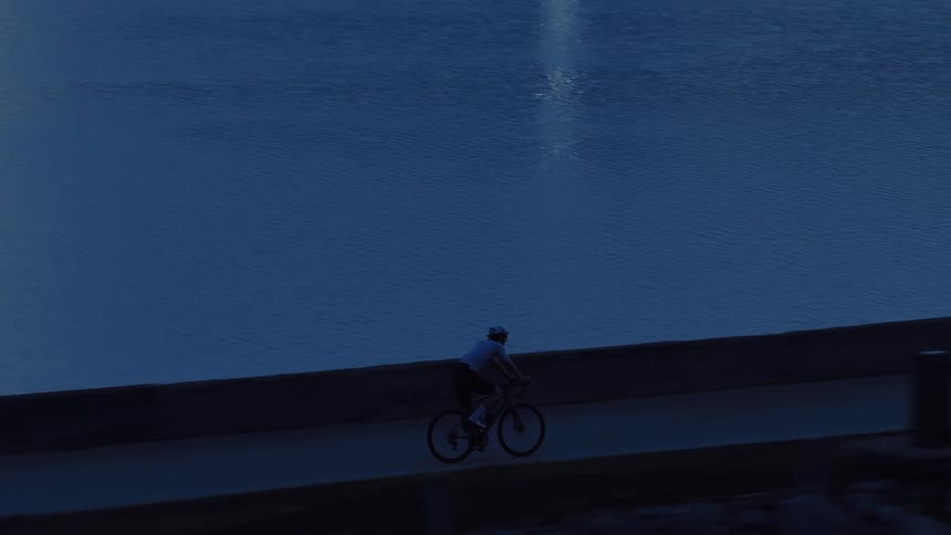 An aerial view follows a determined cyclist training at night along a dark, quiet lakeside road