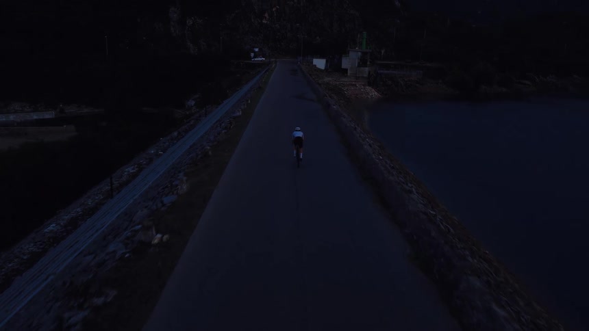 An aerial view shows a lone cyclist riding on a remote road across a dam in the dark of early morning