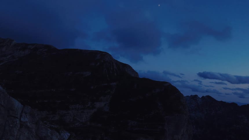 A slow aerial pullback reveals a crescent moon in a deep blue sky over a silhouetted mountain range at dusk