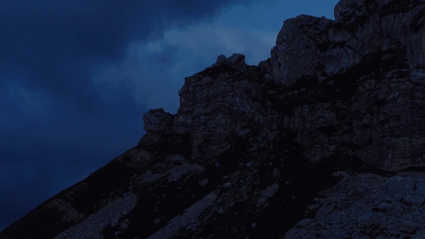 An imposing rocky mountain peak is revealed against a deep blue and cloudy sky during the blue hour