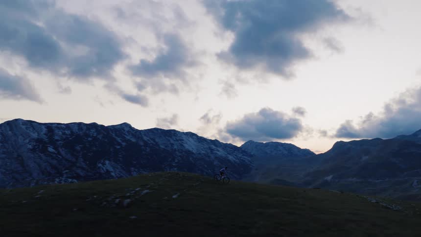 A lone mountain biker rides across a grassy ridge at dusk, silhouetted against a dramatic cloudy sky and a majestic mountain range