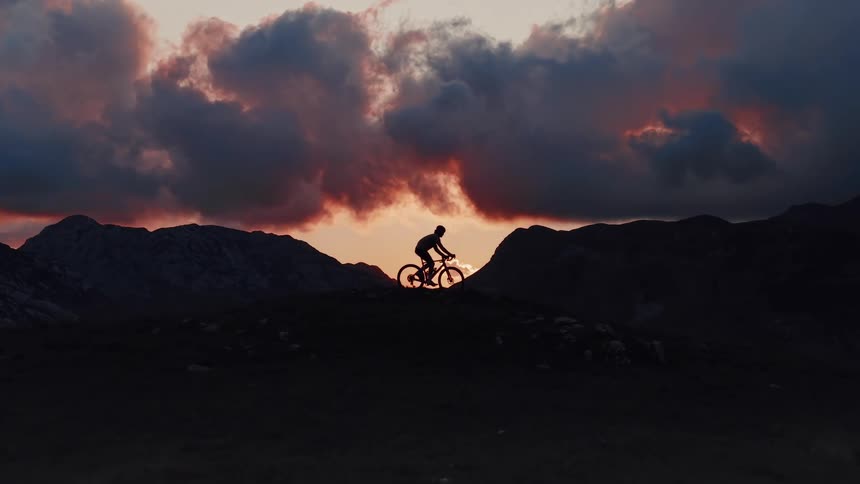 A cyclist rides across a mountain ridge, silhouetted against a dramatic and beautiful sunset sky