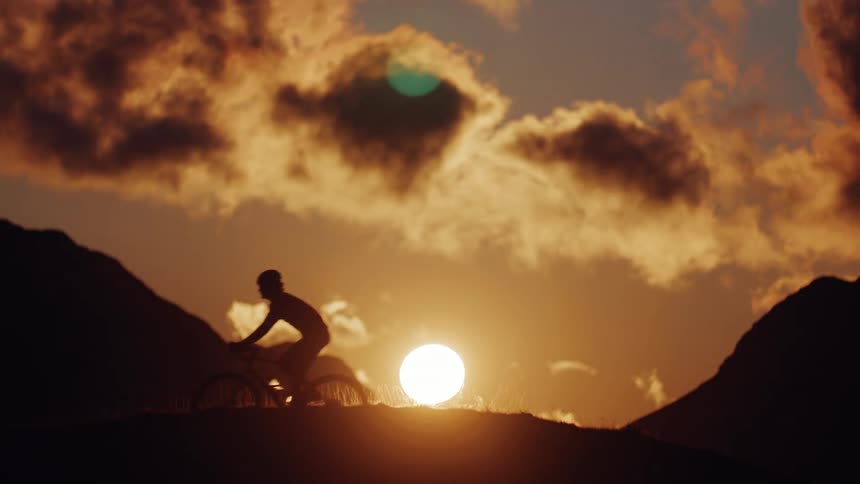 A silhouetted cyclist rides across a mountain ridge in slow motion during a beautiful golden sunset